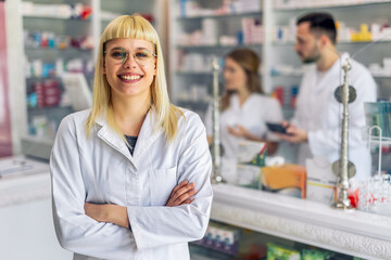 Smiling portrait of young, beautiful female pharmacist that is standing at the cash desk. Other pharmacists working in the background.