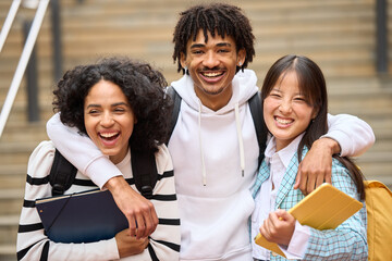 Wall Mural - Three young students are smiling and hugging each other while holding books and looking at camera