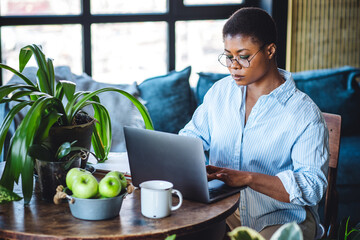 Young attractive African American woman nutritionist is working on laptop in a beautiful green space, home or modern office. Healthy food, apples, water. Video consultation, conference, presentation