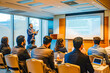 © VisualProduction - A South Asian entrepreneur passionately delivers a presentation on a large screen to a captivated audience in a conference room, bathed in the warm glow of the setting sun outside.