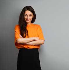 Wall Mural - Beautiful young serious smiling business woman in bright casual orange t-shirt looking with folded arms on grey studio background on empty copy space. Closeup