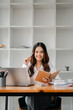 © Satori Studio - A woman is sitting at a desk with a laptop and a notebook. She is smiling and holding a pen. The scene suggests a productive and focused work environment