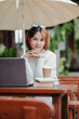 © Satori Studio - A woman is sitting at a table with a laptop and a cup of coffee. She is wearing sunglasses and a white shirt. The scene is set outdoors under an umbrella