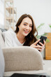© Satori Studio - A woman is sitting on a couch and smiling while holding a cell phone. Concept of relaxation and leisure, as the woman is enjoying her time on the couch