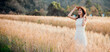 © Satori Studio - A woman is standing in a field of tall grass, wearing a white dress and a straw hat. She is smiling and looking up at the sky. Concept of freedom and relaxation