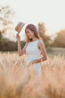 © Satori Studio - A woman is standing in a field of tall grass, wearing a straw hat and a white dress. She is smiling and holding the hat in her hand. Concept of freedom and joy