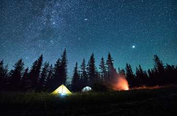 Wall Mural - Night camping under sky full of stars and Milky way in the mountains. Starry sky over illuminated tourist tents on hills near forest. Warm light from campfire at dark night.