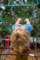 Wall Mural - The child is hanging upside down on the playground equipment. The outdoor playground for children in summer park. Kid play on playground under the tree. Excited kid doing rock climbing.