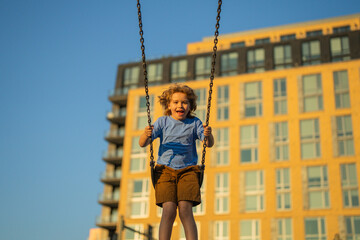 Wall Mural - Excited kid swinging on chain swing on city kids playground. Swing ride. Cute child having fun on a swing on summer sky background. Blonde little boy swings at kid playground. Child swinging high.