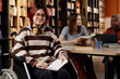 © AnnaStills - Red-haired Asian girl with disability sitting in wheelchair in modern college library looking at camera