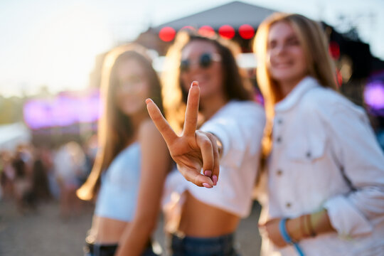 Group of female friends enjoy summer music festival on beach. Smiling girls in trendy outfits dance, laugh, show peace sign. Partygoers celebrate, have fun in sunset light at seaside concert event.