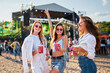 © artiemedvedev - Girls toast with cold drinks at beach music fest in summer, smiling, wearing sunglasses, with stage, people in background. Celebration, fun, youth lifestyle. Females enjoying party, ocean nearby.