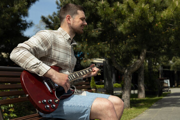 Wall Mural - Attractive young guy playing guitar while sitting on a bench