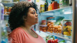 © vetrana - Curvy plus-size woman standing in front of open refrigerator, contemplating food options. She is looking at the food items inside the refrigerator while making a decision on what to eat