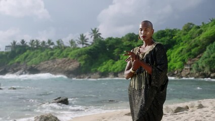 Wall Mural - Elegant gay black man in luxury gown poses on scenic ocean beach. Gender fluid ethnic fashion model in long posh dress and accessories looks at camera stands with sophisticated posture slow motion