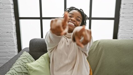 Poster - Cheerful young black woman with dreadlocks casually sitting on a cozy sofa at home, confidently pointing a finger at you and the camera, radiating positivity and a very heartwarming smile!