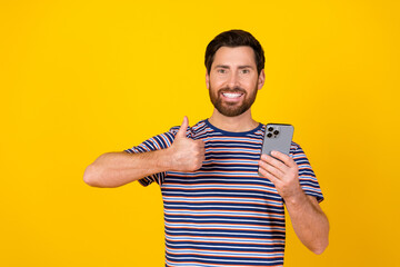 Canvas Print - Photo of satisfied man with beard dressed striped t-shirt hold smartphone showing thumb up isolated on vibrant yellow color background