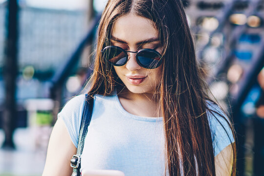 Stylish female skater with long brunette hair and sunglasses holding srateboard in hand while chatting online with followers on smartphone via 4G internet.Female with backpack messaging on cellular
