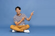 © comzeal - Young Asian man smiling and pointing to empty copy space and sitting on floor isolated on blue background