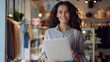 © Johannes - Smiling young woman holding a laptop and looking at the camera standing against a small modern fashion store interior, generative ai