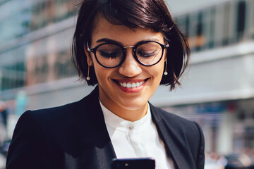 Wall Mural - Smiling female owner reading funny message on smartphone while standing on street near traffic.Successful lawyer in elegant wear received good news on phone with takeaway cup near copy space area