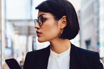 Wall Mural - Serious female trader in eyewear walking on street of megalopolis with tasty coffee in hands.Confident businesswoman in stylish formal wear looking away holding mobile telephone and coffee to go