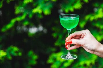 A close-up of a hand holding a glass with a green beverage against a blurred background of lush foliage