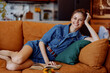 © SHOTPRIME STUDIO - Happy woman relaxing on couch with coffee and book in stylish denim dress in cozy living room setting