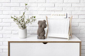 Canvas Print - Blooming branches in vase, statuette and magazine holder on chest of drawers near white brick wall