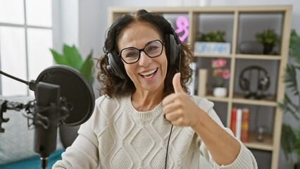 Canvas Print - Cheerful middle age hispanic woman, beaming in approval, gives a cool thumb up sign in radio studio. showcasing success, she’s all smiles using microphone, headphones on.