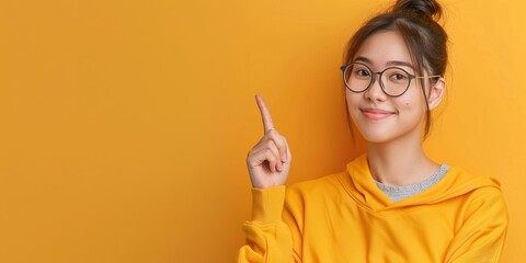 young woman with long wavy brown hair and round glasses pointing with index finger while wearing yel