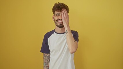 Wall Mural - Confident young hispanic man standing against an isolated yellow background, covering one eye with hand, flashing a smiling face