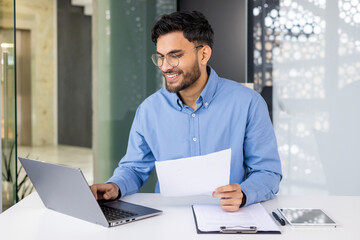 Poster - Smiling businessman working on laptop and holding paper while sitting in modern office