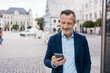 © contrastwerkstatt - Mature businessman in blue suit checking his phone while standing in a modern urban environment with historic buildings.