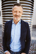 © contrastwerkstatt - A confident senior man in a blue blazer, standing outdoors and smiling at the camera. Business professional with gray hair and beard.