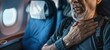 © Panuma - A man experiencing discomfort while sitting in an airplane seat during a flight, suggesting a possible health issue or difficult travel experience.