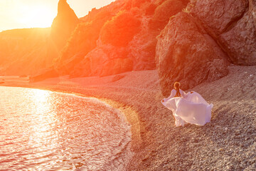 Wall Mural - Mysterious woman silhouette long hair walks on the beach ocean water, sea nymph wind listens to the wave. Throws up a long white dress, a divine sunset. Artistic photo from the back without a face