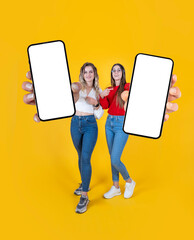 Showing mobile phone with empty white screen, full body length young two women best friends hold show close up to camera smartphone mock up. Yellow studio background. Copy space. New application.