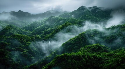 Wall Mural - Lush forested mountain range shrouded in mist, with waterfalls cascading among the greenery on a cloudy day.