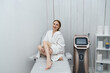 © Anton Pentegov - young beautiful caucasian smiling woman woman in laser epilation cabinet preparing to beauty procedures, sitting on couch and applying cream on leg