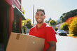 © HNJ - A smiling delivery man in red polo shirt carrying cardboard box on the street
