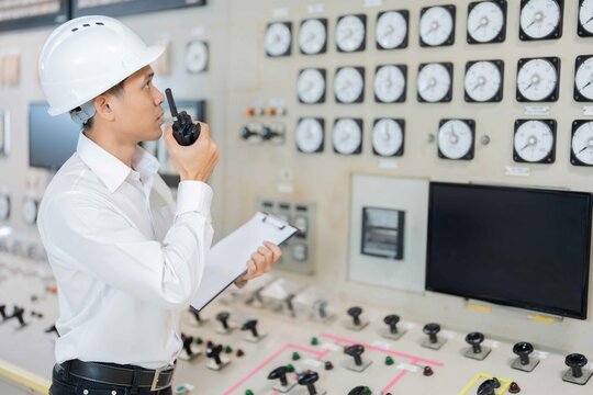 Asian male engineer working on the complex work of a power plant and checking machine circuit boards in a control room holds a list of notes and a radio to check energy production efficiency.