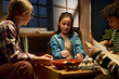 © pressmaster - Smiling pretty girl with dice in hand sitting by table in front of her friends and looking at board while playing tabletop game