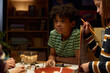 © pressmaster - Cute African American kid in striped t-shirt sitting by table with game board and looking at one of players while choosing roles