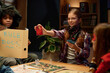 © pressmaster - Cute smiling girl in eyeglasses showing one of her cards to friends while standing by table with game board and character figurines