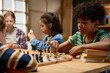 © pressmaster - Clever African American schoolboy moving chess piece on board while sitting by table in front of his friend and playing with him