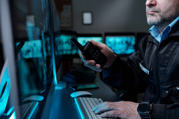 Wall Mural - Cropped shot of mature male security guard with walkie-talkie in hand typing on computer keyboard and looking at screen by workplace