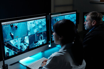 Two intercultural security guards in uniform sitting by workplace in front of computer monitors and observing cctv video on screens