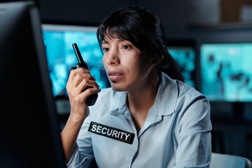 Wall Mural - Young serious Hispanic woman in uniform of security guard sitting in front of computer screen and reporting situation on walkie-talkie