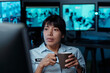 © pressmaster - Young Hispanic female officer working in surveillance room holding cup of coffee and watching cctv video on computer screen